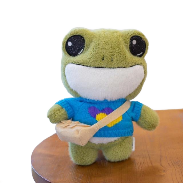 Adorable plush frog toy with a charming design, featuring a small bag and decorative details, placed on a wooden surface.