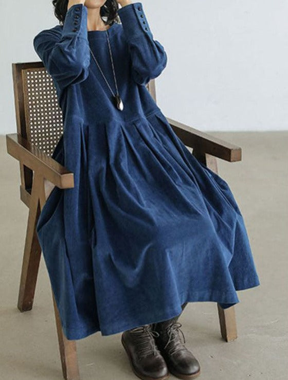 A woman sitting on a wooden chair wearing a vintage midi dress with an elegant design, styled with ankle boots and a long necklace.