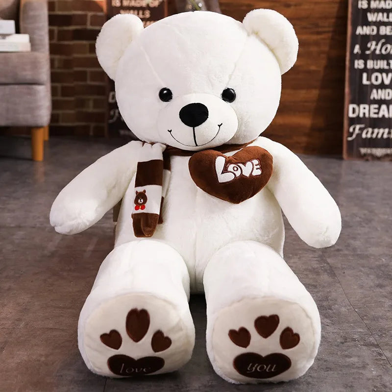 A plush bear toy featuring a heart-shaped patch with "Love" embroidered, wearing a matching scarf with adorable paw print details on its feet, placed on a cozy indoor setting.
