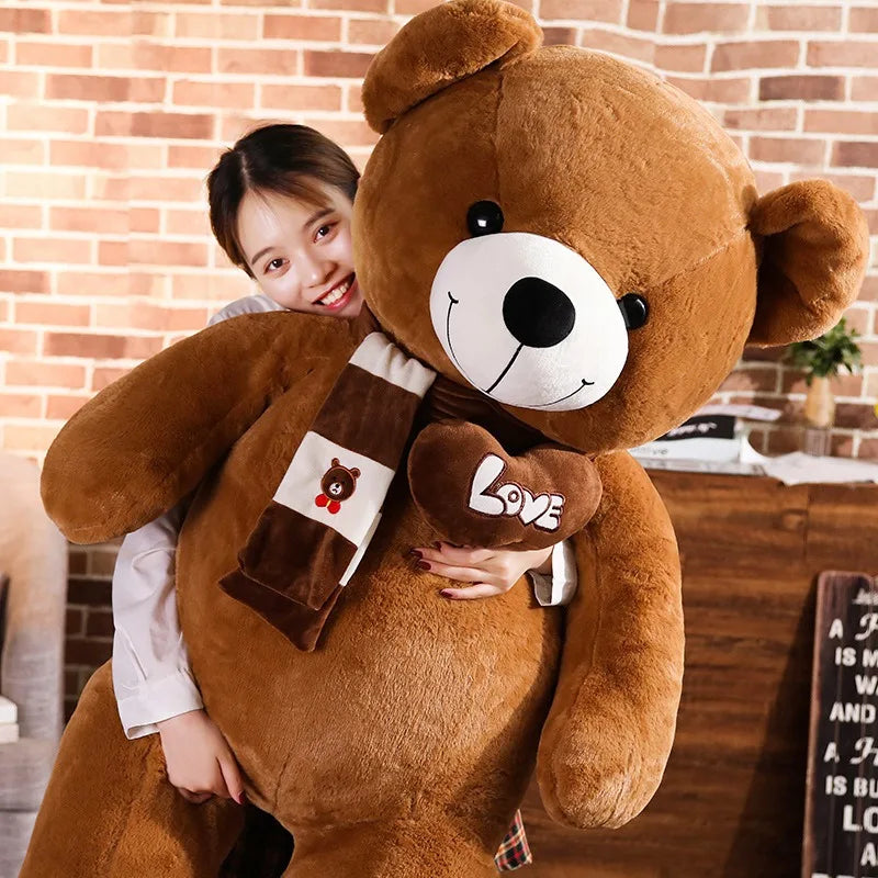 A plush bear toy featuring a heart-shaped patch with "Love" embroidered, wearing a matching scarf with adorable paw print details on its feet, placed on a cozy indoor setting.