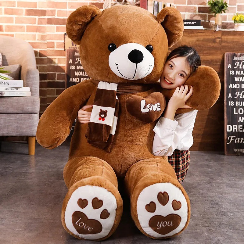 A plush bear toy featuring a heart-shaped patch with "Love" embroidered, wearing a matching scarf with adorable paw print details on its feet, placed on a cozy indoor setting.