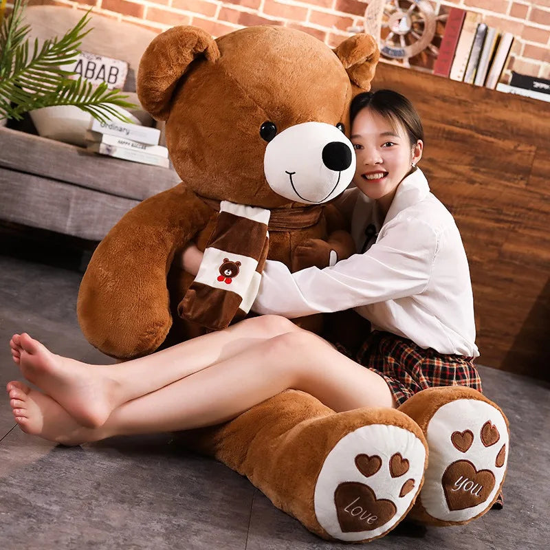 A plush bear toy featuring a heart-shaped patch with "Love" embroidered, wearing a matching scarf with adorable paw print details on its feet, placed on a cozy indoor setting.