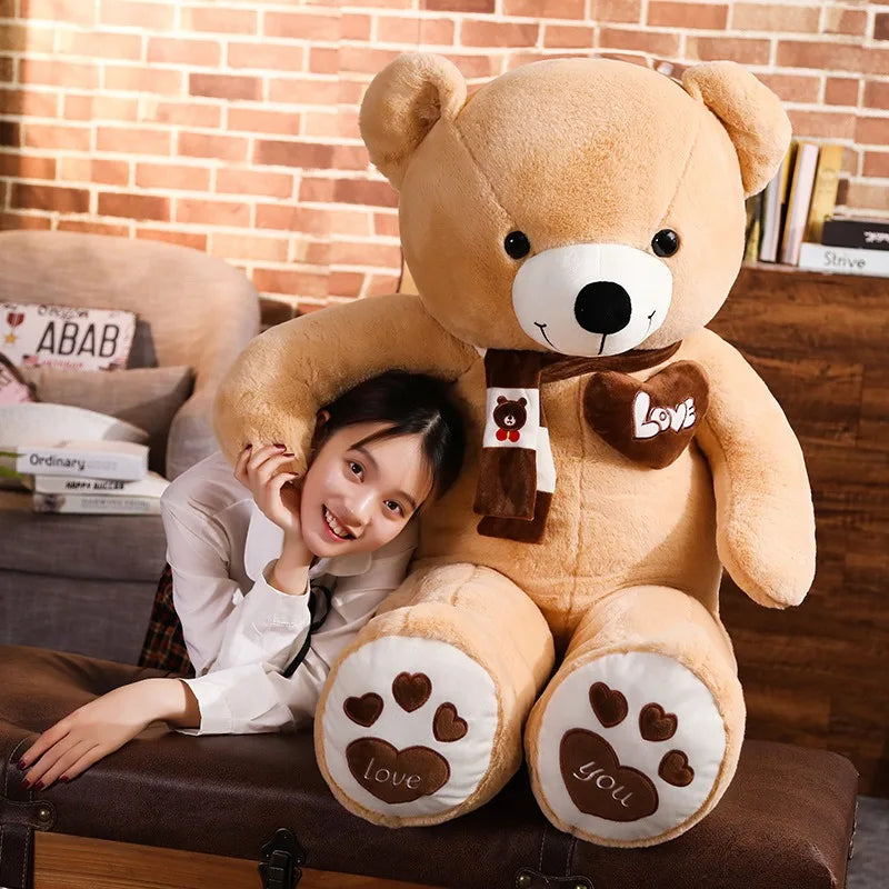 A plush bear toy featuring a heart-shaped patch with "Love" embroidered, wearing a matching scarf with adorable paw print details on its feet, placed on a cozy indoor setting.