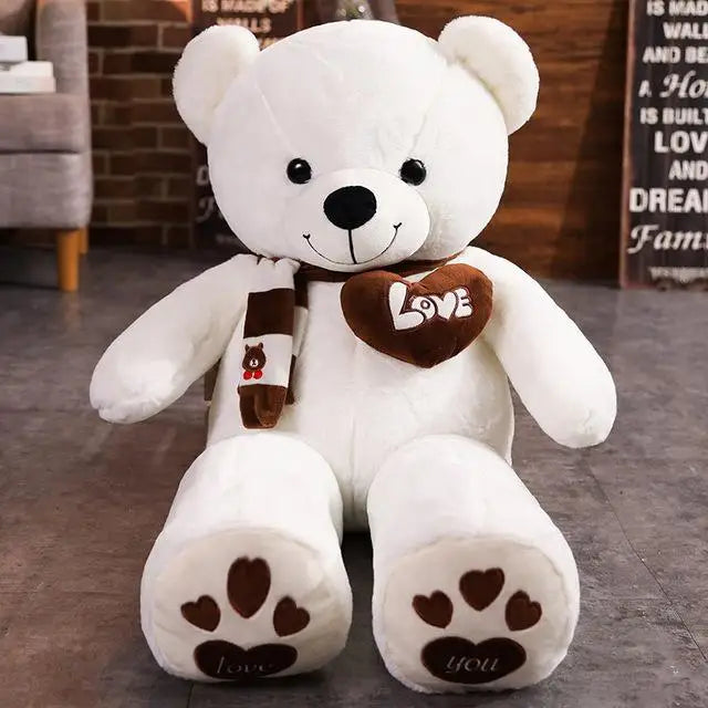 A plush bear toy featuring a heart-shaped patch with "Love" embroidered, wearing a matching scarf with adorable paw print details on its feet, placed on a cozy indoor setting.