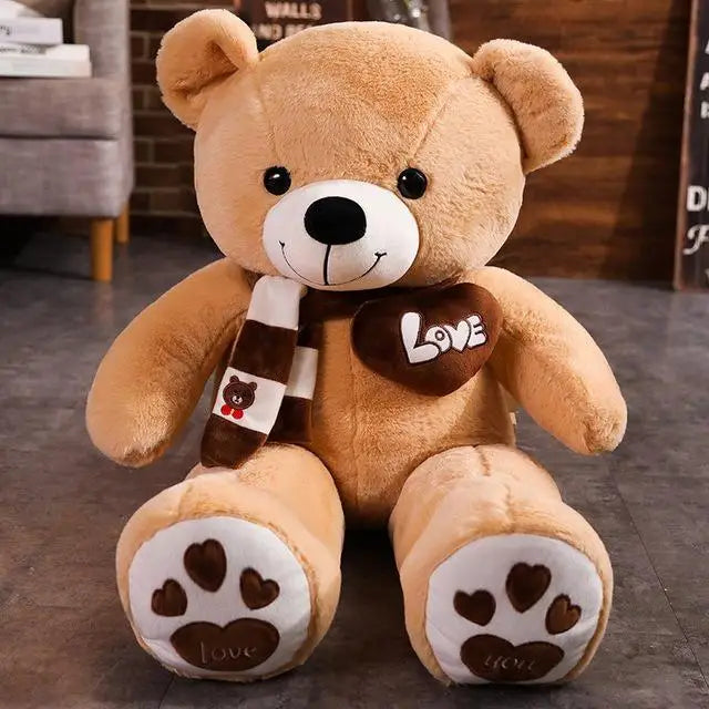 A plush bear toy featuring a heart-shaped patch with "Love" embroidered, wearing a matching scarf with adorable paw print details on its feet, placed on a cozy indoor setting.