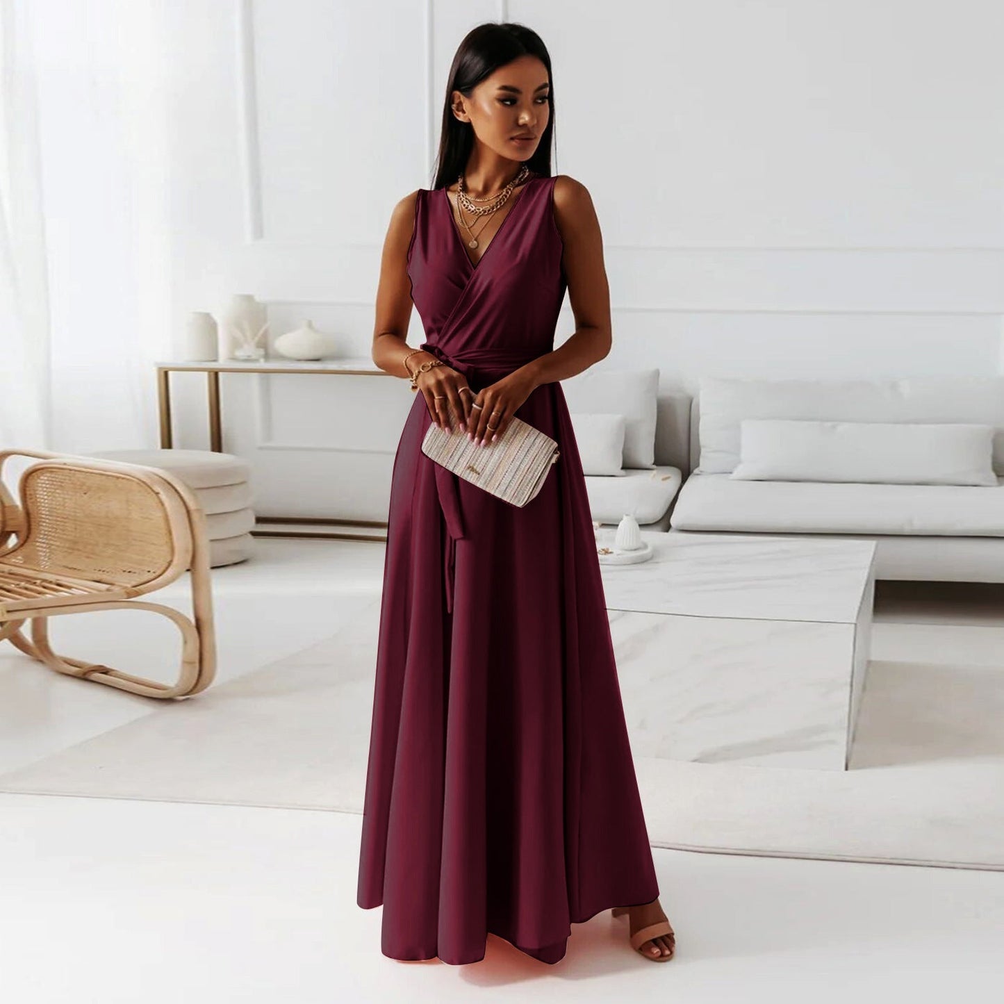 A woman elegantly dressed in a wrap maxi dress featuring a flowing design, styled with minimal accessories, holding a clutch bag, and posing gracefully in a modern living space.
