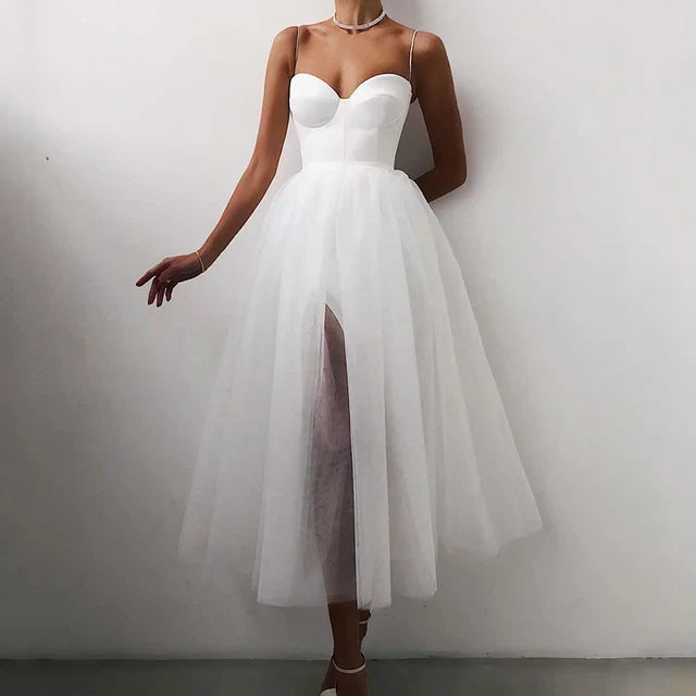 A woman wearing an elegant midi dress with a flowy design, featuring a fitted bodice and a sheer layered skirt, styled with heels and a delicate necklace.
