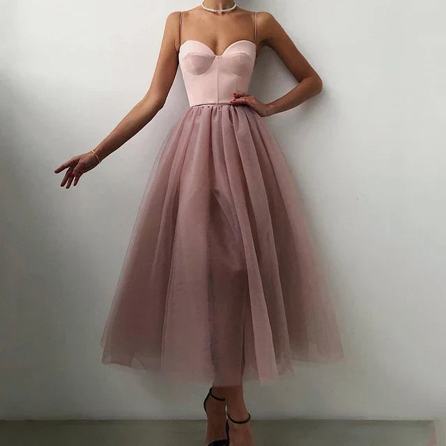 A woman wearing an elegant midi dress with a flowy design, featuring a fitted bodice and a sheer layered skirt, styled with heels and a delicate necklace.