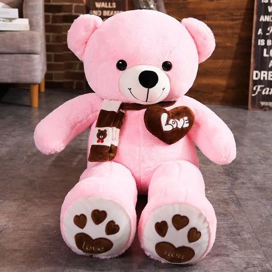 A plush bear toy featuring a heart-shaped patch with "Love" embroidered, wearing a matching scarf with adorable paw print details on its feet, placed on a cozy indoor setting.