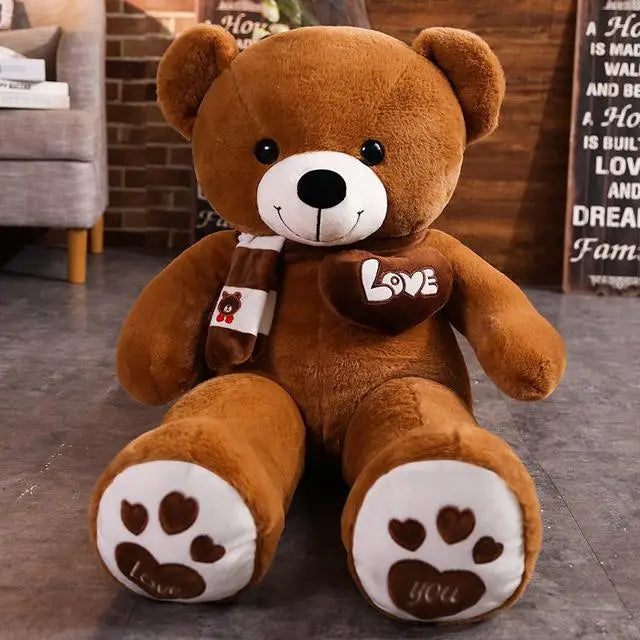 A plush bear toy featuring a heart-shaped patch with "Love" embroidered, wearing a matching scarf with adorable paw print details on its feet, placed on a cozy indoor setting.