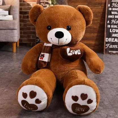 A plush bear toy featuring a heart-shaped patch with "Love" embroidered, wearing a matching scarf with adorable paw print details on its feet, placed on a cozy indoor setting.
