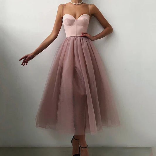 A woman wearing an elegant midi dress with a flowy design, featuring a fitted bodice and a sheer layered skirt, styled with heels and a delicate necklace.